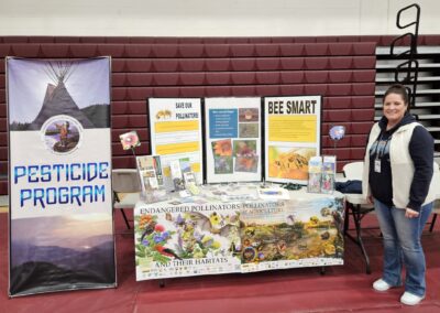 Woman stands in front of table with informational posters and pamphlets with titles "pesticide program", "Bee smart", and "save our pollinators"